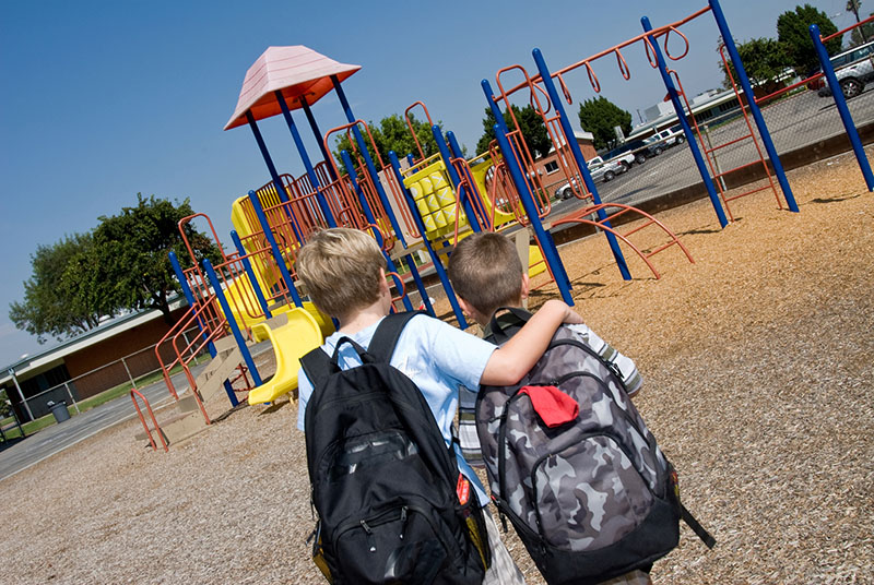 Two children with backpacks sharing a side hug in a school playground