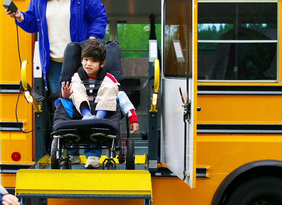 A child with disability sitting in a wheelchair at a school bus door with a student supervisor behind him