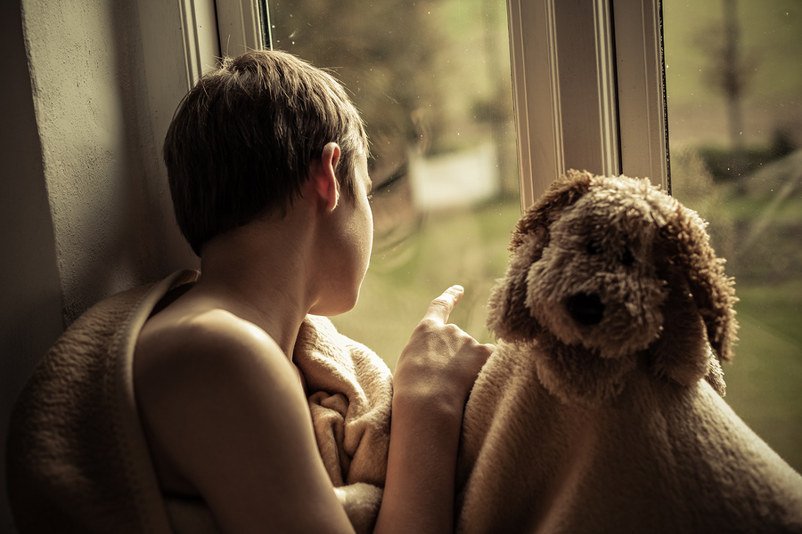 A sad child sitting by the window with a blanket and a stuffed animal