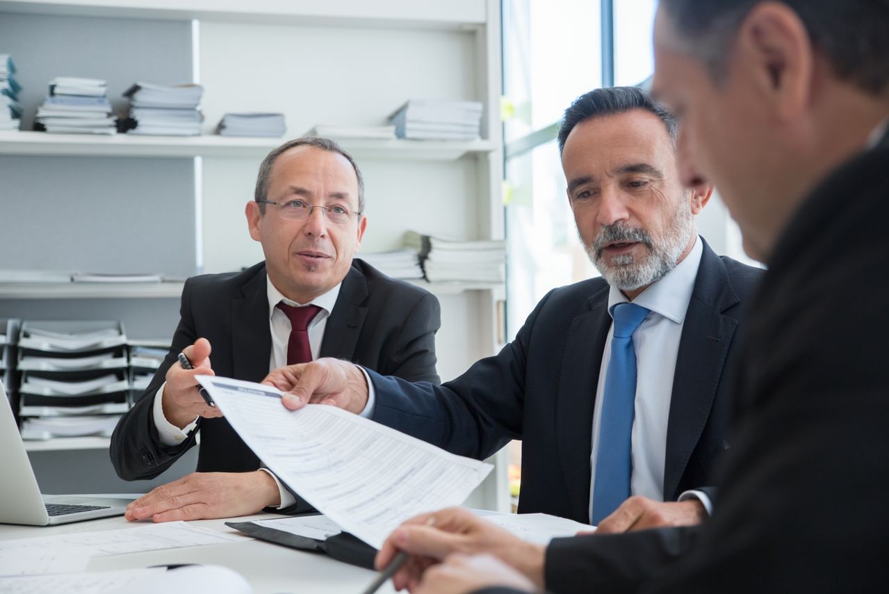 Three men in black suits reviewing documents in an office